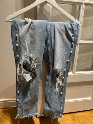 Ljusa jeans med pärlor och slitningar - Säljer ett par ljusblå jeans med raka ben, råa kanter och stora slitningar vid knäna. Jeansen har pärldetaljer längs sidorna och klassisk femficksdesign. Perfekta för dig som gillar en edgy och unik stil.