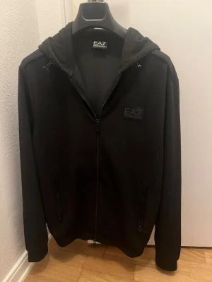 Emporio Armani zip hoodie - Stilren EA7 zip hoodie med luva i väldigt skönt material med en logga som smälter in med hoodien på ett snyggt sätt. Storlek L. Mycket bra skick, har bara hängt i garderoben. Har ej sett denna hoodie i Sverige eftersom den är från London, rätt så sällsynt! 