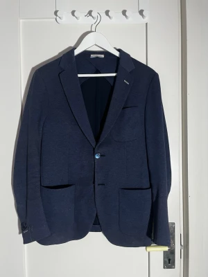 Marinblå kavaj från Gant Rugger - Säljer en marinblå kavaj från Gant Rugger med två knappar framtill, stora fickor och klassisk krage. Materialet är bomull och modellen är 'unconstructed' vilket ger en avslappnad känsla. Perfekt för dig som vill ha en stilren men chill look.