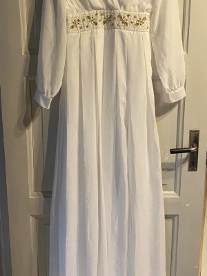 Vit långklänning från Nelly - Elegant vit långklänning från Nelly med v-ringad omlottdesign och långa ärmar. Midjan pryds av ett brett band med glasstenar. Tillverkad i ett lätt, flytande polyestertyg som ger ett drömmigt intryck.