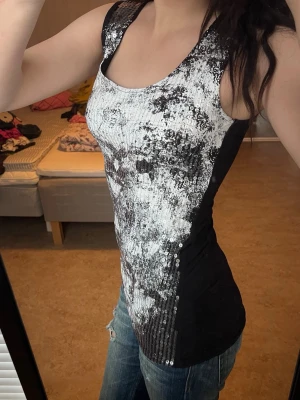 Svart och vit paljettlinnetopp - Säljer en snygg ärmlös linne-topp i svart och vitt med glittriga paljetter över hela framsidan. Toppen har ett abstrakt mönster och är figurnära med breda axelband. Perfekt för dig som vill sticka ut lite extra.