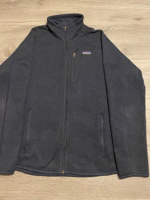 Mörkgrå fleece från Patagonia - Exklusiv Patagonia fleece i storlek M i perfekt skick – 10/10. Endast varsamt använd. Ett plagg som känns och ser ut som nytt.  Patagonia står för premiumkvalitet, tidlös design och komfort i toppklass. Denna fleece är både mjuk och värmande, perfekt för en stilren vardagslook eller som ett elegant mellanlager under kyligare dagar.  Ett genomtänkt val för dig som uppskattar kvalitet, hållbarhet och stil. Missa inte chansen att göra ett riktigt bra köp!