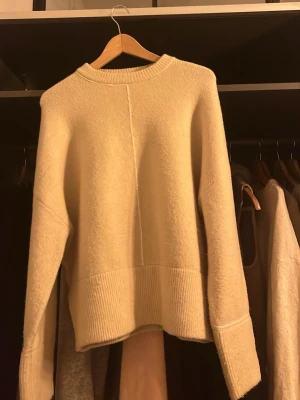 Beige stickad tröja med ribbade detaljer - Mysig beige stickad tröja med breda ribbade muddar vid ärmslut och nederkant. Tröjan har en rak passform och en markerad söm framtill som ger en cool touch. Perfekt för lager på lager under kyliga dagar.