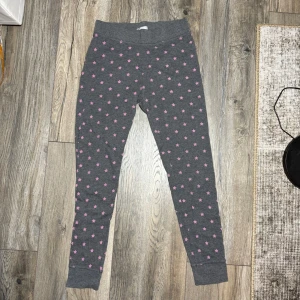 Grå leggings med rosa stjärnor - Mysiga grå leggings från Cubus i storlek S, täckta med små rosa stjärnor. De har bred resår i midjan och muddar vid bensluten för extra komfort. Perfekta för chill dagar hemma eller som ett färgglatt inslag i vardagslooken.