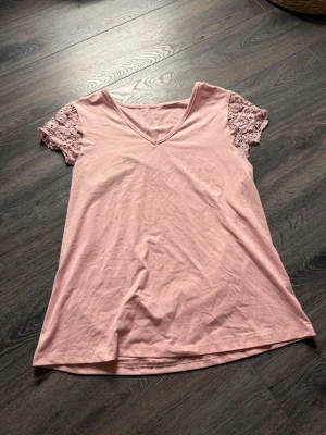 Ljusrosa topp med spetsärmar - Supersöt ljusrosa topp med v-ringning och korta spetsärmar. Toppen har en enkel, rak passform och är gjord i mjuk bomull. Spetsdetaljerna på ärmarna ger en romantisk vibe och gör den extra fin till både jeans och kjol.