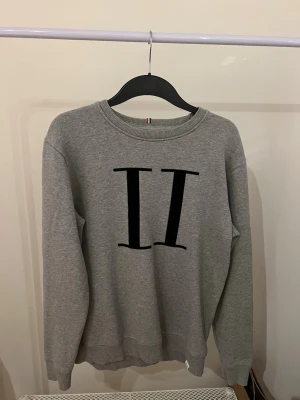 Grå tröja från Les Deux - Säljer en grå sweatshirt från Les Deux med stor svart 'II'-logga på bröstet. Tröjan har rund halsringning, ribbade muddar och är tillverkad i mjuk bomullsmix. Perfekt för en chill och stilren look. 