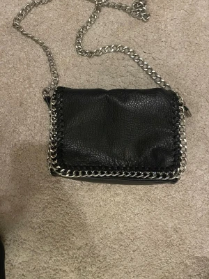 Svart axelväska från Tiamo med kedja - Säljer en svart axelväska från Tiamo i skinnimitation med grov silverfärgad kedja som axelband och detaljer runt kanterna. Väskan har dragkedja både på insidan och utsidan samt magnetknäppning. Perfekt storlek för det viktigaste och ger en cool vibe.