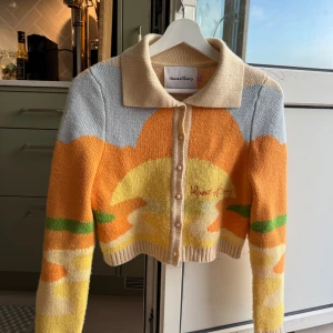 House of Sunny kofta med solnedgång - Superfin kofta från House of Sunny med solnedgångsmotiv i orange, gul, beige, grön och ljusblå. Croppad modell med krage, knappar framtill och långa ärmar. Perfekt statement-plagg för dig som gillar färg och unika mönster. Kostade 1500kr ny.