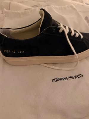 Common Projects Achilles Low Suede - Snygga svarta sneakers från Common Projects i mocka med vit sula och vita snören. Minimalistisk design med guldfärgad numrering på sidan och diskret branding. Tillverkade i Italien och levereras med skopåsen plus två extra skosnören. Storlek 42. Helt nya och oanvända. Nypris 390 Euro (ca 4200kr)  Finns att hämta på Östermalm. Snabb affär = bra pris. Pris går att diskuteras 