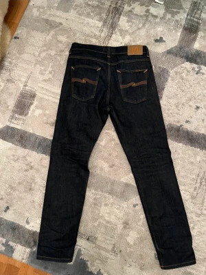 Nudie Jeans Steady Eddie II - Säljer ett par mörkblå jeans från Nudie Jeans med klassiska kontrastsömmar och snygga bakfickor med den ikoniska vågdetaljen. Modellen är Steady Eddie II med normal midja och tillverkad i slitstarkt jeansmaterial. Perfekta för dig som gillar en stilren look. Använda ett fåtal gånger och jeansen tyg är så kallat Selvage jeans. Nypris 1800kr mitt pris 600kr