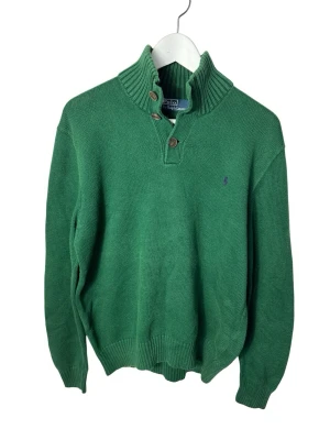 Grön stickad tröja Polo Ralph Lauren - Grön stickad tröja från Polo Ralph Lauren med ribbad hög krage och två knappar framtill. Klassisk broderad logga på bröstet och långa ärmar. Perfekt till höst och vinter för en chill men ändå snygg look.