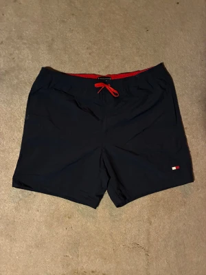 Marinblå badshorts Tommy Hilfiger - Säljer ett par marinblå badshorts från Tommy Hilfiger med röd dragsko och röd insida i midjan. Klassisk logga framtill och en stor flaggdetalj bak på fickan. Perfekta för stranden eller poolen.