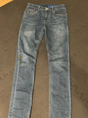 Levi's blå  jeans - Säljer ett par klassiska blå Levi's jeans i skinny fit med fem fickor och snygga slitningar. Jeansen har orange kontrastsömmar och en tydlig Levi's-logga på insidan av linningen. Perfekta till sneakers eller boots. Lite smutsiga men jag kan tvätta om det behövs stl 30/33