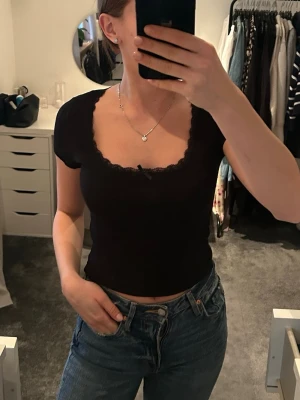 Svart croppad t-shirt med spetskant - Säljer en svart croppad topp med korta ärmar och u-ringning. Toppen har en söt spetskant runt urringningen och en liten rosett framtill. Materialet är mjukt och stretchigt.