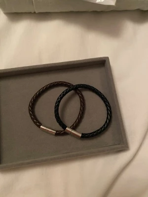 Två flätade läderarmband svart & brun - Säljer två stilrena armband i flätat läder, ett i svart och ett i brunt. Båda har en enkel magnetlåsning i silverfärgad metall. Perfekta accessoarer för en clean och modern look.