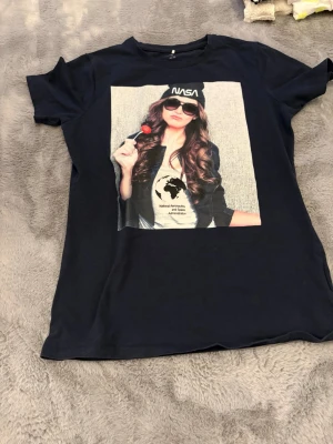 Svart t-shirt med tryck från Name It - Svart t-shirt från Name It med coolt tryck av en tjej som håller en klubba och har en NASA-mössa. Gjord i mjuk ekologisk bomull och har en klassisk passform med korta ärmar. Perfekt för dig som gillar statement-plagg och snygga prints.