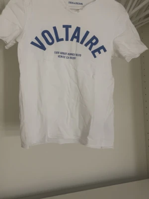 Vit t-shirt från Zadig & Voltaire - Vit t-shirt från Zadig & Voltaire med blå text VOLTAIRE framtill. Klassisk passform och rund hals. Tillverkad i 100% bomull för en skön och luftig känsla. Perfekt till jeans eller shorts för en clean och enkel look.