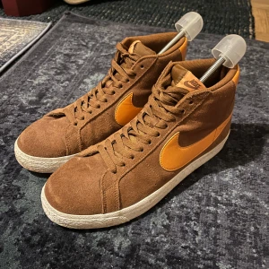 Bruna Nike SB Blazer Mid sneakers - Säljer ett par bruna Nike SB Blazer Mid sneakers i mocka med orange swoosh och detaljer. Skorna har vit sula, snörning och klassisk hög siluett. Perfekta för dig som gillar retrovibbar och streetstyle. Passar till både skate och vardagsoutfits. Knappt använda 