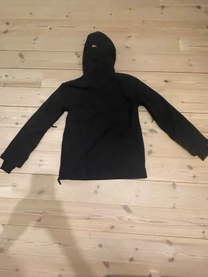 Svart/blå skidjacka från Quiksilver - Säljer en svart vindjacka från Quiksilver med huva och en stor blå ficka framtill. Jackan har en orange half zip-dragkedja och detaljer i olivgrönt. Perfekt för dig som vill ha en stilren men ändå färgglad jacka. Funkar bra som skidjacka. Bara att höra av sig vid frågor👍