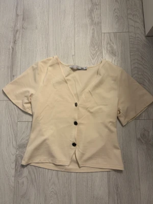 Beige kortärmad blus från 157 - Snygg beige blus från 157 med v-ringning och svarta knappar framtill. Toppen har korta ärmar och är lätt croppad, perfekt till jeans eller kjol. Tillverkad i mjuk bomull som känns skön mot huden.