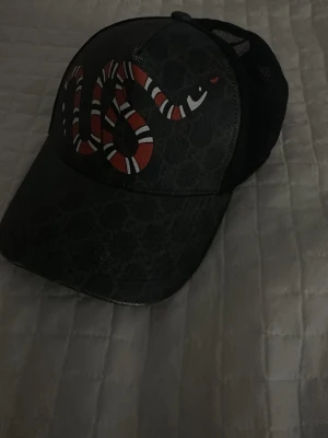 Svart keps med ormtryck  - Säljer en svart keps med ett fett ormtryck i rött, vitt och svart på framsidan. Kepsen har mesh på sidorna och justerbart band bak med röd och grön rand. Perfekt accessoar för att lyfta din outfit. Självklart inte äkta 