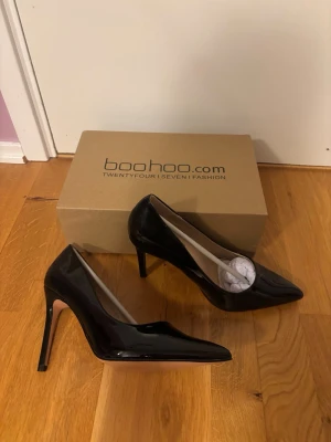 Svarta lackpumps från Boohoo - Snygga svarta pumps i glansigt lackmaterial från Boohoo. Skorna har spetsig tå och hög klack som ger en elegant siluett. Perfekta för dig som vill ha en klassisk och stilren look med lite attityd.