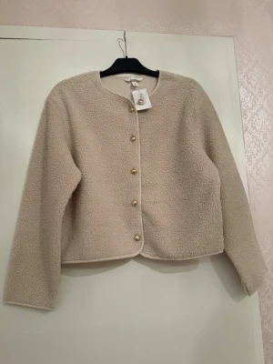 Beige teddyjacka med guldknappar - Såldes för 299kr. Aldrig använd. Supermjuk beige teddyjacka från H&M med rund halsringning och snygga guldknappar framtill. Jackan har lång ärm och en rak, lite boxig passform. Perfekt för dig som gillar en clean och cozy look.