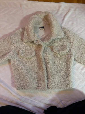 Beige teddyjacka från Gina Tricot - Supermysig beige teddyjacka från Gina Tricot i storlek M. Jackan har bröstfickor, krage och stängs med tryckknappar. Perfekt för dig som vill ha en varm och trendig look i höst och vinter. Lite nopprig inuti men går att enkelt ta bort