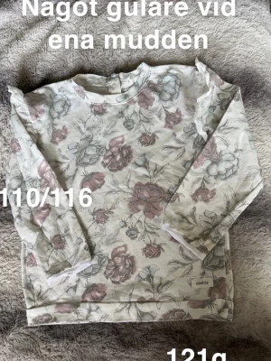 Blommig långärmad tröja Newbie - Supersöt långärmad tröja från Newbie med blommigt mönster i dova toner av rosa, grått och grönt. Tröjan har volangdetaljer på axlarna och rund halsringning med knäppning bak. Perfekt för vår! 🌸 Något gulare vid ena mudden. Storlek 110/116 
