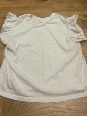 Vit blus med volangärm från Zara - Supersöt vit blus från Zara med korta volangärmar och rund hals. Tillverkad i mjuk bomull som känns skön mot huden. Perfekt för vår och sommar när du vill ha en enkel men ändå stylish look.