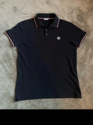 Svart Moncler piké med rödvit kant - Säljer en mörkblå pikétröja från Moncler med klassisk krage och knappar framtill. Tröjan har röda och vita ränder på krage och ärmslut samt Moncler-logga broderad på bröstet. Tillverkad i bomull för en skön och luftig känsla. Självklart äkta och köpt i cph förra året! Skit bra skick osså