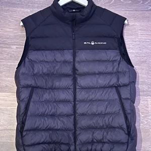 Svart dunväst från Sail Racing - Snygg svart dunväst från Sail Racing med hög krage och dragkedja framtill. Västen har två praktiska fickor med dragkedja och logga på bröstet samt en logga i nacken. Perfekt för lager-på-lager och stilren look.