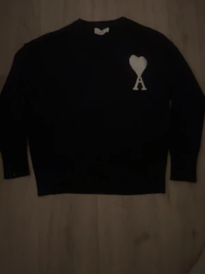 Ami Paris  - Säljer en navy blå stickad tröja från AMI Paris med vit hjärtdetalj och bokstaven A på bröstet. Tröjan har lång ärm och klassisk passform, perfekt för dig som gillar stilrena plagg med en twist.