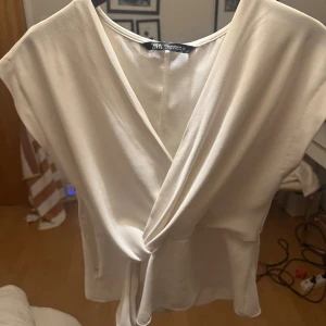 Vit omlottopp från Zara XS - Säljer en stilren vit omlottopp från Zara i storlek XS. Toppen har en snygg v-ringning, knyt i sidan och ärmlös design. Materialet känns mjukt och har ett lätt glansigt utseende som ger en lyxig känsla. Perfekt att matcha med jeans eller kjol.