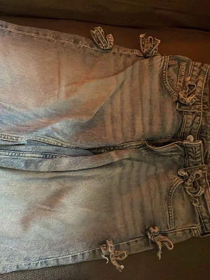 Ljusblå jeans med rosettdetaljer! - Unika ljusblå jeans med flera snörningsdetaljer längs sidorna och vid fickorna. Klassisk femficksmodell med dragkedja och knapp. Jeansen har en tvättad look och raka ben. Perfekta för dig som vill sticka ut med coola detaljer.