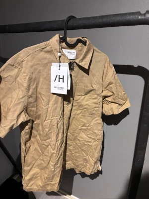 Beige half zip skjorta från Selected Homme - Kortärmad beige skjorta från Selected Homme i bomull med relaxed fit och krage. Skjortan har en bröstficka och en snygg half zip-dragkedja framtill. Perfekt till jeans eller shorts för en chill look.