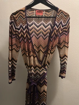 Mönstrad omlottklänning Missoni x Lindex - Säljer en superfin omlottklänning från Missoni x Lindex med ikoniskt zigzagmönster i lila, beige, svart och guld. Klänningen har trekvartsärm, v-ringning och knyt i midjan. 