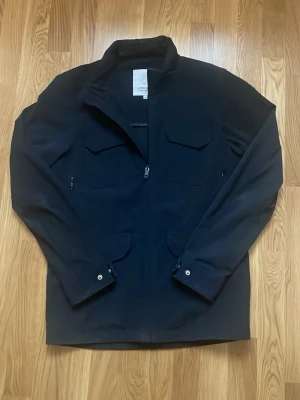 Marinblå field jacket från Elvine - storlek S. Jackan har fyra fickor med lock framtill, dragkedja hela vägen och justerbara ärmslut med knapp. Enkel och clean design som funkar till allt. 