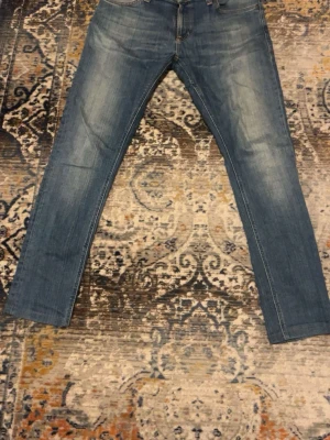 Blå jeans från Nudie Jeans - Säljer ett par klassiska blå jeans från Nudie Jeans med snyggt slitna detaljer och kontrastsömmar. Modellen har raka ben, fem fickor och Nudie Jeans ikoniska broderi på bakfickorna. Perfekta för dig som gillar en tidlös och avslappnad look. Lagade vid skrevet.