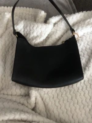 Svart axelväska  -  svart axelväska i skinnmaterial. Väskan har guldfärgade metalldetaljer och dragkedja upptill. Den följer med en liten ring som går att sätta dit och av. Väskan är lite sönder på sidan som på sista bild. Kommer gå ner i priset mkt sen men väljer nu 21kr då de kanske blir billigare eftersom de är fri frakt nu.