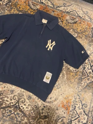 Mörkblå Yankees henley från Starter - Säljer en mörkblå henley från Starter Cooperstown Collection med New York Yankees-logga på bröstet. Skjortan har korta ärmar, klassisk krage och half zip framtill. Tillverkad i mjuk bomull och har en vintage-inspirerad patch nertill.