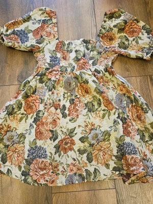 Blommig kortklänning med puffärm - Supersöt kortklänning med stora puffärmar och ett romantiskt blommönster i beige, rost, grön och blå. Klänningen har smock i ryggen för skön passform och är gjord i ett tjockare bomullsliknande tyg. Perfekt för dig som gillar vintage vibes och statement-ärmar.