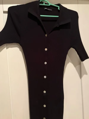 Svart ribbad långklänning från Zara - Säljer en svart ribbad långklänning från Zara med korta ärmar. Den är aldrig använd.  Klänningen har knappar hela vägen ner och är figurnära i modellen. Den formar kroppen väldigt fint !!!