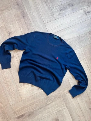Mörkblå tröja från Polo Ralph Lauren - Klassisk mörkblå tröja från Polo Ralph Lauren i 100% pima bomull. Tröjan har rund halsringning, och den ikoniska röda loggan broderad på bröstet. Perfekt till jeans eller chinos för en clean och stilren look.