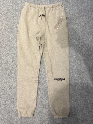 Essentials mjukisbyxor med snörning - Säljer ett par gråa  mjukisbyxor från Essentials Fear of God i storlek M. Byxorna har resår i midjan, långa snören framtill och muddar vid bensluten. Tillverkade i mjukt bomullsmaterial och har loggan tryckt på benet. Perfekta för chill dagar.