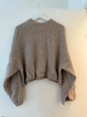 Beige/brun stickad croppad tröja Gina Tricot - Säljer en beige/brun, grovstickad croppad tröja från Gina Tricot. Tröjan har breda, långa ärmar och en hög ribbad krage. Perfekt för lager på lager under höst och vinter. Supermjuk och oversized fit för en chill vibe.