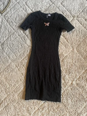 Svart kort klänning från H&M Divided - Säljer en svart kort klänning från H&M Divided i storlek XS. Klänningen har en tight passform, korta ärmar och ett smockat tyg som ger en cool struktur. På bröstet sitter en broderad fjäril i rosa toner som detalj.