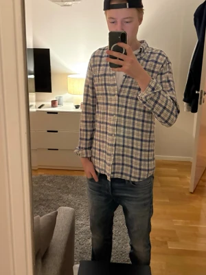 Blåvit rutig skjorta med knappar - Säljer en blåvit rutig skjorta med knappar framtill och klassisk krage. Skjortan har lång ärm och är tillverkad i ett mjukt bomullstyg. Perfekt att bära öppen över en t-shirt för en avslappnad look.