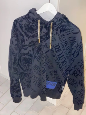 Versace Jeans Couture Hoodie – Svart - Snygg och exklusiv hoodie från Versace Jeans Couture i svart ton-i-ton barockmönster. Stilren men samtidigt iögonfallande design med diskret logotryck i tyget och guldfärgade kedjedetaljer i huvan som ger en lyxig känsla. Mjuk och skön kvalitet, perfekt både till vardags och för en mer uppklädd streetwear-look.  
