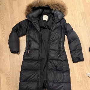 Svart lång dunjacka från Moncler - Säljer en svart lång dunjacka från Moncler med stor huva och fluffig pälsdetalj. Jackan har dragkedja, två fickor fram och ett brett bälte med Moncler-logga i ryggen. Perfekt för kalla dagar och riktigt snygg siluett.